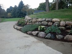 there is a large rock wall in the middle of this driveway with flowers growing on it