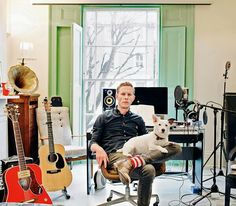 a man sitting in a chair with a dog on his lap next to guitars and other musical equipment