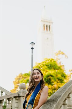 a woman wearing a blue and yellow sash standing on a stone wall with a clock tower in the background