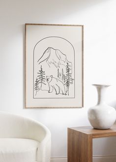 a white vase sitting on top of a wooden table next to a wall mounted drawing