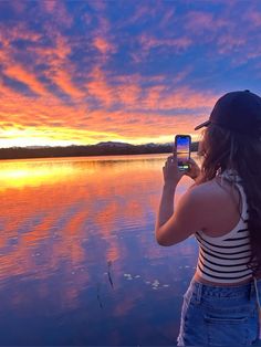 a woman taking a photo with her cell phone at sunset on the water's edge