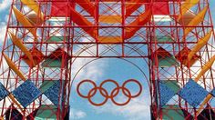 an olympic sign is seen through the scaffolding on top of a structure in front of a cloudy blue sky