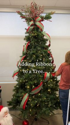 a woman standing next to a christmas tree with red and green ribbons on it's top