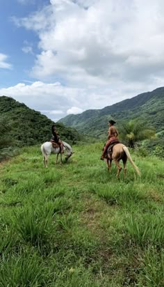 two people are riding horses in the grass on a hill side with mountains in the background