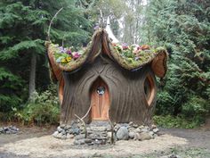 a house made out of logs and grass with plants growing on the roof, surrounded by trees