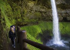 a woman standing in front of a waterfall next to a lush green forest covered hillside