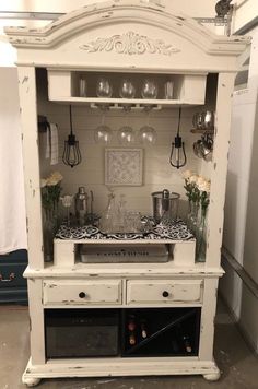 an old white china cabinet with wine glasses on top and flowers in the bottom drawer