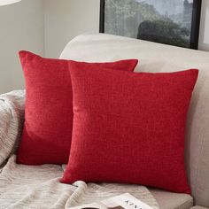 two red pillows sitting on top of a white couch next to a book and lamp