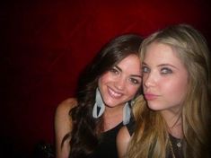 two beautiful young women standing next to each other in front of a red wall and smiling at the camera