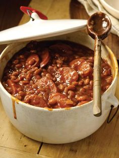 a pot filled with beans and sausages on top of a wooden table next to a spoon