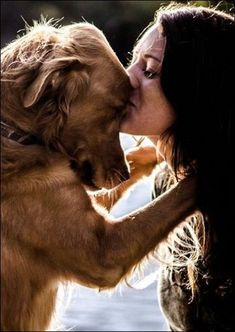 a woman kissing a dog on the nose with water in the back ground behind her