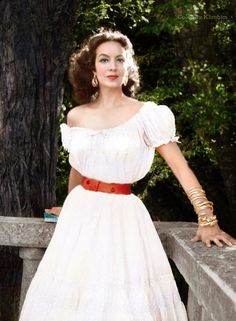 a woman in a white dress standing on a stone wall next to a tree and wearing gold bracelets