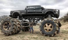 two men stand next to large tires on a truck that is lifted in the air