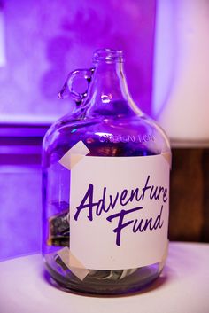 a purple glass jug filled with coins sitting on top of a white table next to a sign that says adventure fund