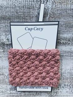 a pink crocheted cup cozy on top of a white wooden table with the words cup cozy written above it