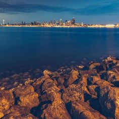 the city skyline is seen in the distance from some rocks on the shore at night