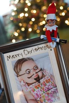 an elf is holding a christmas card in front of a framed photo with the caption merry christmas