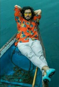 a man with long hair and glasses sitting in a boat on the water, wearing blue shoes