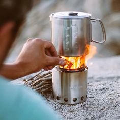 a person lighting a campfire in a stainless steel mug on the ground with sticks