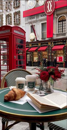 an open book, coffee cup and croissant on a table in front of a red phone booth