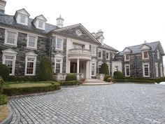 a large stone house with many windows and bushes on the front lawn, surrounded by cobblestones