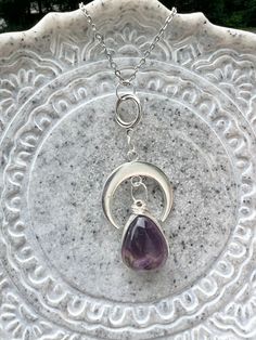 This statement necklace features a gorgeous large amethyst gemstone. The amethyst is wire wrapped by hand with silver wire and bezel set on a silver pear shaped base. The stone hangs from a brilliant silver moon adding a celestial element to the pendant. The moon is suspended from a dainty antique silver twist hoop. The deep purple stone is domed on top and flat on the back. Many dark and light shades of purple are displayed throughout the stone. The color variations were created by the minerals that grew with the amethyst in nature. The stunning pendant is 63.3 mm long and is attached to a beautiful silver stainless steel chain with a lobster clasp. Amethyst is a grounding, protective, calm stone that encourages balanced energy, love of the divine, and selflessness. * Information for meta Adjustable Purple Moon Phase Jewelry, Spiritual Crescent Wire Wrapped Necklaces, Silver Crescent Necklace With Natural Stones, Bohemian Wire Wrapped Amethyst Necklace, Bohemian Amethyst Moon Charm Jewelry, Nickel-free Amethyst Necklace For Healing, Handmade Silver Crescent Crystal Necklace, Bohemian Silver Teardrop Pendant Crystal Necklace, Purple Bohemian Jewelry With Moon Charm