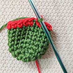 two knitting needles are next to a green and red crocheted object on a white surface