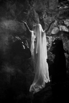 a woman dressed in white standing next to a rock wall with her arms outstretched and head down