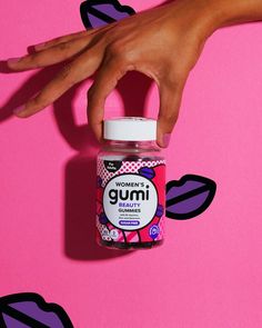 a woman's hand reaching for a gumi bottle on a pink background with black and purple shapes