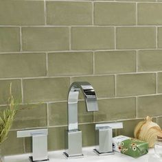 two faucets on a sink in front of a green brick wall and vase with flowers