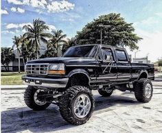 a large black truck parked on top of a parking lot next to trees and palm trees