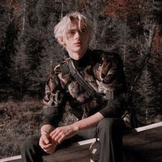 a young man sitting on top of a wooden bench in front of a forest filled with trees