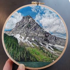 someone is holding up a hand embroidered mountain scene with trees and clouds in the background