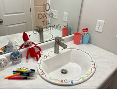 an elf is sitting in the bathroom sink with markers and crayons on the counter