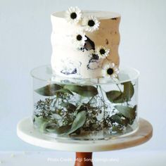 a three tiered cake with white frosting and flowers on the top is sitting on a wooden stand