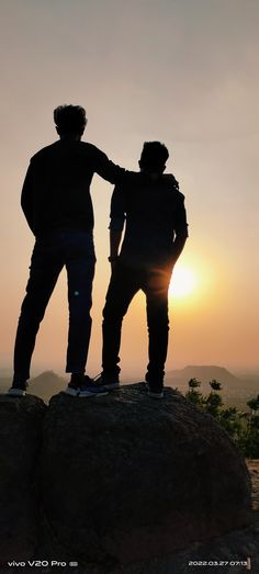 two people standing on top of a rock with the sun setting in the distance behind them