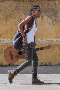 a man walking down the street with a guitar in his hand and a bag on his shoulder