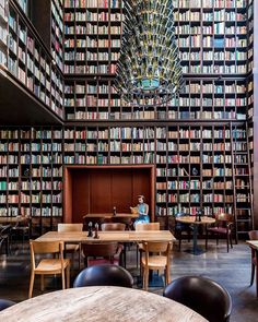 a library filled with lots of books next to wooden tables and chairs in front of a chandelier