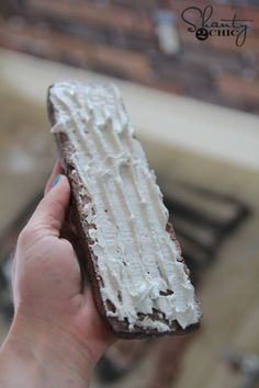 a hand holding a piece of cake with white frosting on it and a brick wall in the background