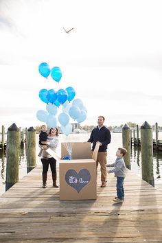 a family standing on a dock with balloons floating in the air and a cardboard box that says it's us