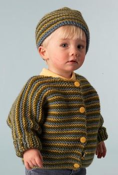 a young boy wearing a knitted sweater and hat with buttons on the collar, standing in front of a gray background