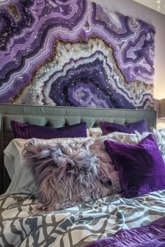 a bed with purple and white pillows on it next to a wall hanging above the headboard