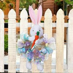 a white bunny with a carrot in its mouth hanging on a fence post and wearing a colorful bow