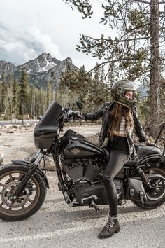 a woman sitting on top of a motorcycle in front of some trees and mountain peaks