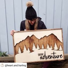 a woman holding up a sign that says see adventure with pine trees and mountains in the background