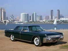 an old green car parked in front of the water and cityscape behind it