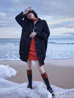 a woman standing on top of a beach next to the ocean wearing rain boots and an overcoat