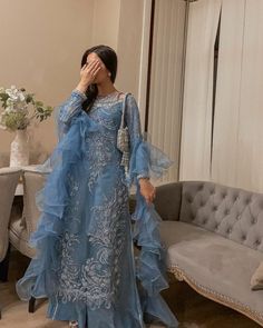 a woman standing in front of a couch wearing a blue dress and holding her hand to her face