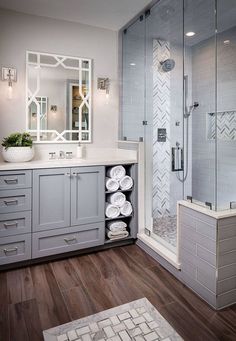 a bathroom with gray cabinets and white towels on the counter top, shower stall, and wooden flooring