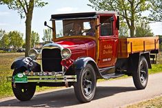 an old red truck driving down a road next to some grass and trees on a sunny day